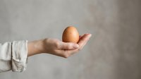 Person with light skin tone holding a brown egg in open palm against neutral gray background, suitable for food, nutrition, cooking, and agricultural marketing materials.