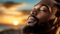 Young African American man with beard in peaceful meditation pose against dramatic sunset sky with glowing sun and clouds. Wellness and mindfulness concept.