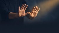 Male hands reaching forward in dramatic lighting with open palms extended in protective or defensive gesture against dark moody background.