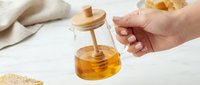 Hand holding glass honey dispenser with wooden dipper drizzling golden honey, natural sweetener for kitchen cooking and baking on white background.