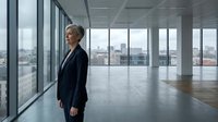 Mature woman in business attire standing in modern empty office space with floor-to-ceiling windows overlooking cityscape, suitable for corporate and professional concepts.
