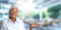 Confident senior woman with glasses and white hair gesturing with open palm against blurred modern interior background.