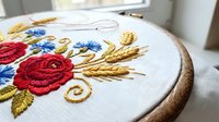 Embroidery hoop with colorful floral design featuring red roses, blue flowers and golden wheat ears on white fabric for needlework and crafting projects.
