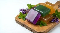 Handmade natural soap bars in purple, green and orange colors with fresh lavender and mint herbs on wooden cutting board for organic skincare and spa treatments.