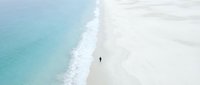 Aerial view of solitary person walking on pristine white sand beach with turquoise ocean waves creating peaceful coastal scenery for travel and vacation concepts.