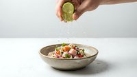 Hand squeezing fresh lime juice over colorful vegetable salad in ceramic bowl on white surface, food preparation and healthy eating concept.