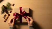 Hands tying red ribbon bow on kraft paper gift box with pine branches, cinnamon sticks and star anise on wooden background for holiday wrapping.