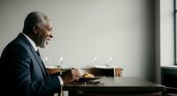 Senior African American male executive in dark business suit dining alone at wooden table with plate of food and glass of water in modern office setting with natural window light.
