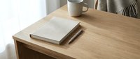 Wooden desk with stacked notebooks and ceramic mug in bright natural light, suitable for workspace and office design projects.