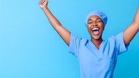 Young African American female healthcare worker in blue scrubs and cap celebrating with arms raised, expressing pure joy and success against blue background.