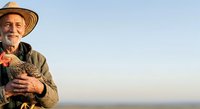 Senior male farmer of Caucasian ethnicity wearing straw hat and work clothes, holding fresh harvested vegetables outdoors at sunrise for agricultural and farming industry use.