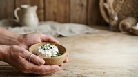 Mature male hands holding wooden bowl with fresh cheese and herbs on rustic wooden table, vintage kitchen setting with ceramic pitcher and rope, suitable for food photography, culinary blogs, and artisanal product marketing.