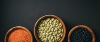 Three wooden bowls filled with legumes and pulses including red lentils, green lentils, and black beans arranged on dark background for cooking and nutrition content.