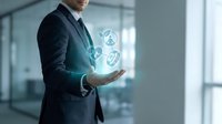 Businessman in dark suit holding glowing digital icons representing technology and innovation in modern office environment with glass windows and blurred background.