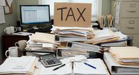 Cluttered office desk with tax documents, calculator, computer and coffee cups during busy accounting season preparation work.
