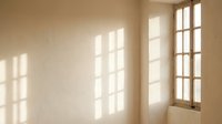 Minimalist empty room interior with natural sunlight streaming through large windows creating geometric shadow patterns on beige textured wall and floor.