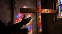 Silhouette of hands raised in prayer inside gothic church with colorful stained glass light reflections on stone walls and wooden cross.