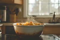 Steaming hot soup with chunks of vegetables in white ceramic bowl on kitchen counter, window light creates cozy atmosphere. Natural food photography.