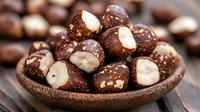 Dark chocolate covered macadamia nuts in wooden bowl on rustic table. Close up view of gourmet confectionery snack with selective focus and natural lighting.
