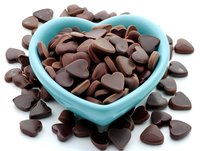 Heart-shaped chocolate pieces in turquoise ceramic bowl, surrounded by scattered chocolate drops on white surface. Sweet romantic food styling for dessert and confectionery.