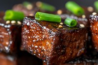Glazed pork belly chunks with caramelized coating and fresh green onion garnish on dark background, extreme close up macro food photography.