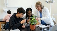Middle-aged Caucasian female teacher in white lab coat instructing diverse teenage students examining potted plant in modern science classroom laboratory setting.