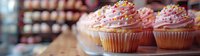 Pink frosted cupcakes with colorful sprinkles arranged on display in bakery, shallow depth of field creates soft background blur. Sweet dessert photography.