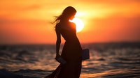 Young woman in flowing dress stands silhouetted against dramatic orange sunset over ocean, holding book while her hair flows in breeze, creating romantic atmosphere.