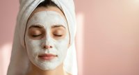 Young woman with white clay facial mask and towel on head relaxing with closed eyes during spa skincare treatment for healthy glowing complexion.