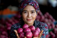 Asian woman vendor in traditional headscarf holding fresh red onions at local market, displaying fresh produce with warm genuine smile, authentic cultural marketplace scene.