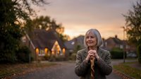 Happy mature blonde woman in beige coat standing outdoors during golden hour sunset in residential neighborhood with warm lighting and peaceful atmosphere.