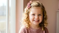 Adorable young Caucasian girl with curly blonde hair and pink bow smiling warmly by window in soft natural light wearing cozy knit sweater at home.