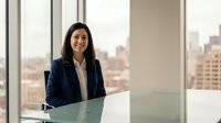Professional Asian businesswoman smiling confidently in modern office with city skyline view, corporate executive at glass desk for business presentations.