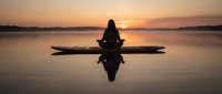 Woman meditating in lotus position on wooden log over calm lake water at golden sunset with perfect reflection creating peaceful zen moment for mindfulness practice.