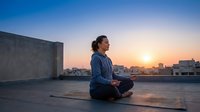 Young Asian woman practicing meditation on urban rooftop at sunset with city skyline background, peaceful mindfulness and wellness lifestyle concept.