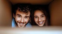 Young Caucasian couple peeking through cardboard box opening with cheerful smiles, moving day concept, relocation and new home lifestyle moment.