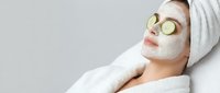 Young woman relaxing with cucumber slices on eyes and white facial mask during spa treatment, wearing towel on head and bathrobe for skincare routine.
