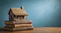 Wooden house model on stack of books about home building and architecture on wooden table against blue background for real estate education.
