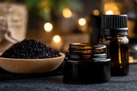 Black seed oil in dark glass jar with wooden bowl of black cumin seeds and essential oil bottles, illuminated by warm bokeh lights, suitable for wellness, aromatherapy and natural beauty product marketing.