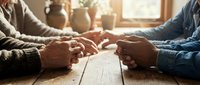Elderly couple holding hands across wooden table in warm sunlight streaming through window creating intimate moment of love and connection in cozy home setting.