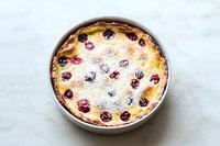 Homemade cherry clafoutis dusted with powdered sugar in white ceramic baking dish on marble surface, traditional French dessert with fresh berries.