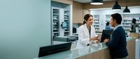 Female pharmacist in white coat consulting with male customer at modern pharmacy counter discussing medication and healthcare services.