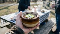 Hands holding ceramic bowl with hot soup outdoors at picnic table with friends gathering in background for camping meal.
