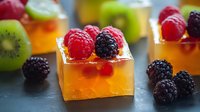 Orange jelly dessert cubes topped with fresh raspberries and blackberries, garnished with kiwi fruit, served on dark surface with natural lighting.