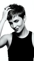 Woman with short dark hair wearing black sleeveless top, hand touching head, smiling at camera in studio portrait against white background for fashion and beauty campaigns.