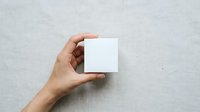 Hand holding blank white square card against textured gray wall background, suitable for branding, business card mockups, and marketing material presentations.