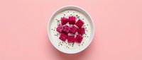 Overhead view of white bowl with creamy yogurt topped with fresh red rose petals and dark seeds on soft pink background, suitable for food and wellness content.