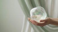 Young adult female with light skin tone holding a transparent glass sphere in cupped hands against soft green and white curtain background, symbolizing care, fragility, and environmental protection.