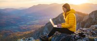 Young woman in yellow jacket working on laptop while sitting on mountain cliff at sunset, overlooking valley landscape for remote work and travel concepts.