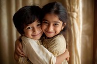 Two young South Asian children embracing and smiling warmly at camera, wearing beige outfits against soft brown curtained background, capturing pure joy and sibling love.